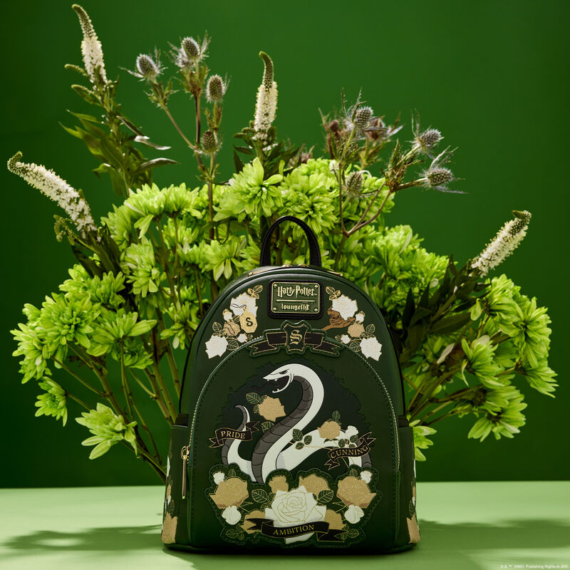 Green mini backpack featuring the Slytherin snake mascot on the front pocket, surrounded by white and pale gold flowers, sitting against a green background in front of a real bouquet of green and white flowers  